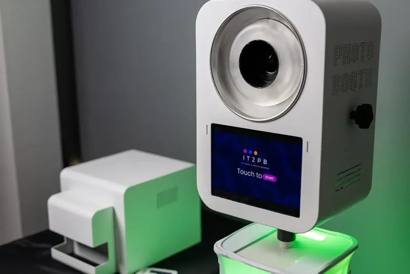 It Takes 2 selfie booth with ring flash and touch screen, next to its dye-sub photo printer, lit with green LED