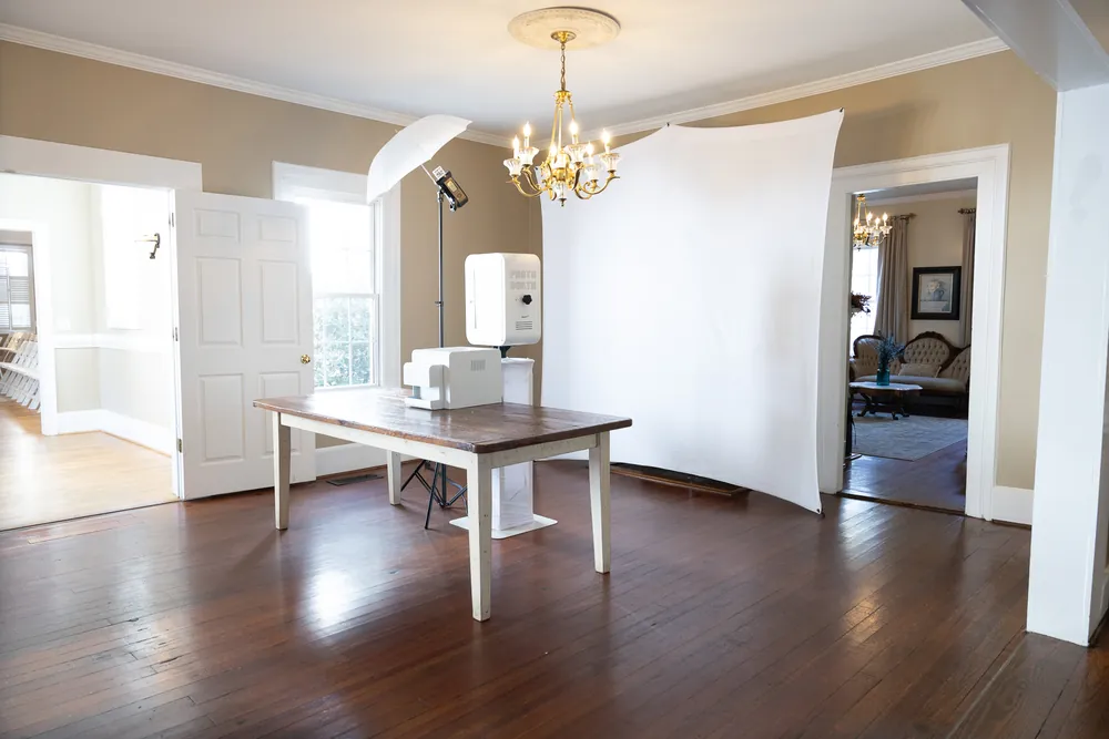 Our standard setup in a venue room: an 8 by 8 white seamless backdrop, the white photo booth on a stand, a studio flash with a white umbrella off to the side, and the printer on a table